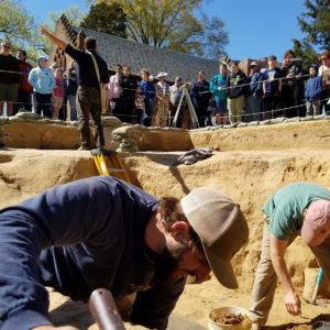 Two archaeologists trowel in a unit while another leads a tour in the background