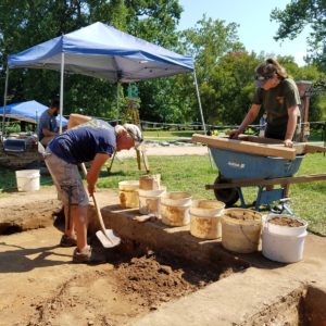 Archaeologist digs with a shovel while another screens artifacts