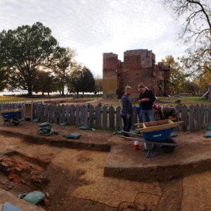 Archaeologists stand in open excavation units in front of house ruins and fence