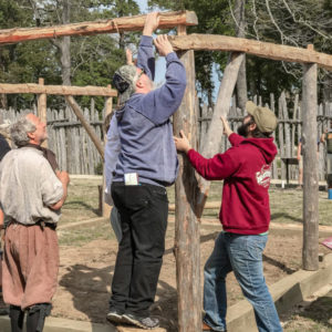 Several men work to fit beams together at the corner of a frame structure