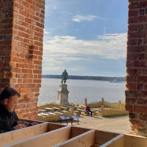 Man screws a wooden platform together inside of a brick tower next to a doorway looking out onto a riverbank