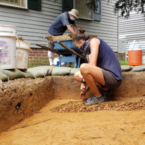 archaeologist trowels in a unit along the side of a house while another screens for artifacts