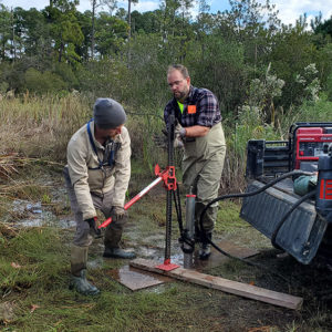 Archaeological Field Technician Gabriel Brown and Dr. David Leslie use a truck jack to extract the vibracore tube from the swamp.