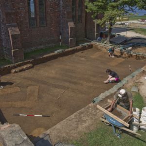 Visitors watch students excavating planting furrows next to brick church