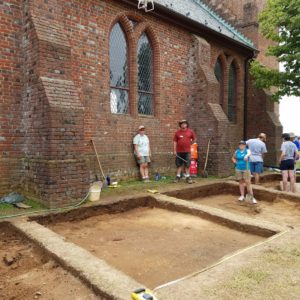 Excavators watch drone fly above excavated units next to brick church