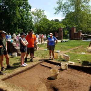 Group of students stand around excavation unit containing brick wall