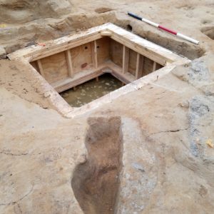 Excavated pit and well lined with wooden siding in the background