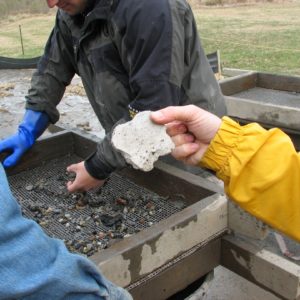 Plaster piece being held up to the camera with view of archaeologists screening artifacts in the background