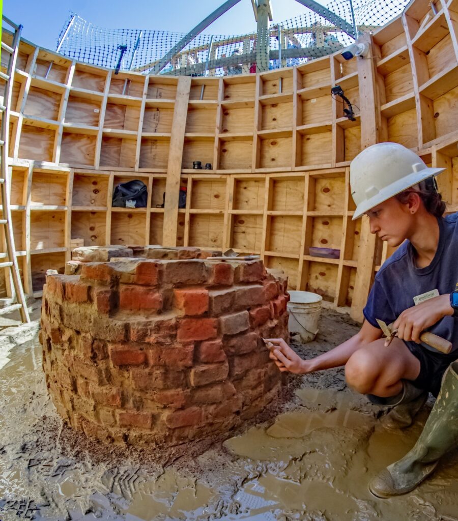 A person in a hard hat uses a small tool to clean mud off of bricks making up the walls of a well. They are surrounded by a structural wall built of wood. The ground is muddy.