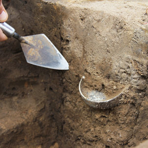 Trowel pointing to half of a shallow round glass dish protruding from an excavation unit wall