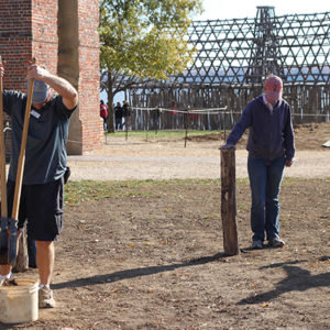 Archaeologist using a large tool to excavate an area while another stands beside a small post