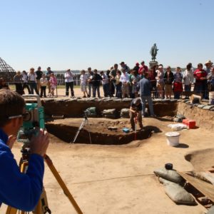 Archaeologist maps feature with a transit while another gives a tour to a crowd in the background