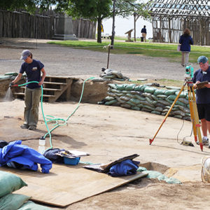 Archaeologist spraying an excavated area with a hose while another records features with a transit