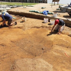 Archaeologists troweling in an excavation area with a reconstructed palisade and a river in the background