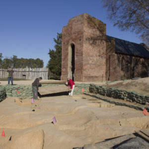 Two men lowering a plywood board to cover an excavation area lined with sandbags in front of a brick church