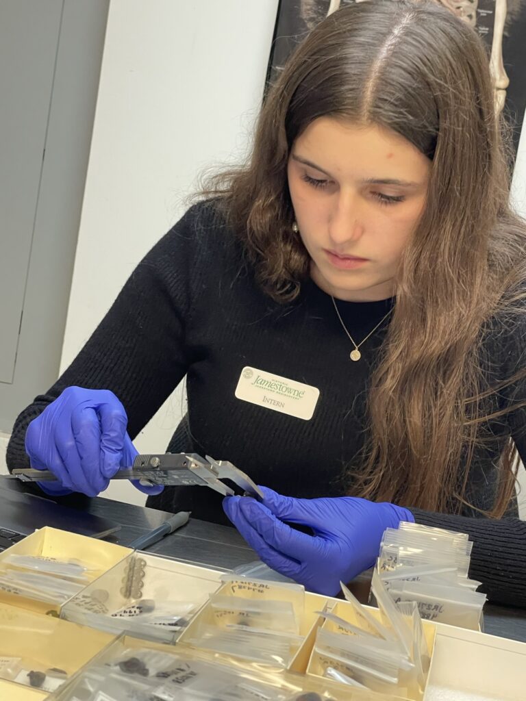 A woman sits at a table with small boxes of artifacts in front of her. She's using a measuring tool to measure a small button in her gloved hand.