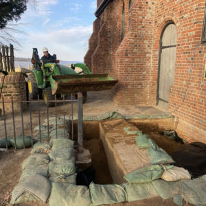 man stands with a shovel next to a man operating a backhoe to fill in two excavation units next to a brick church