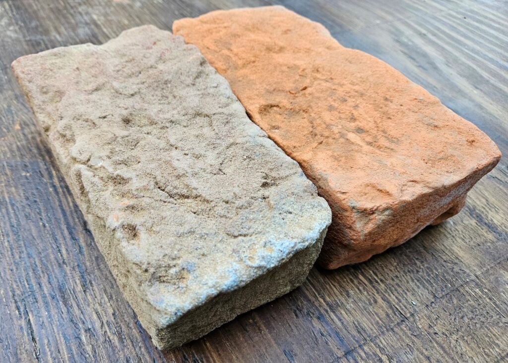 Two bricks side-by-side on a table. One on the left is more tan and has a rough surface. One on the right is more orange and has a smoother surface.