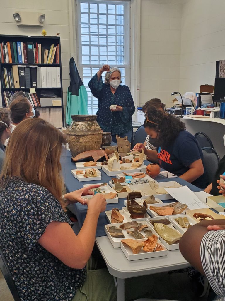 Senior Curator Merry shows off several artifacts excavated at Jamestown that are related to slavery. The group sits around a table covered in boxes of ceramics, glass, and other artifacts. 