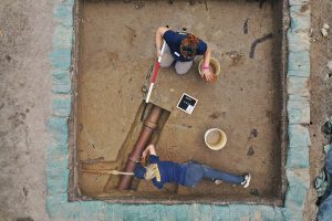 Archaeological Field Technicians Hannah Barch (top) and Katie Griffith excavate around the large drainage pipe and iron water pipe that used to feed a horse trough found in the barracks excavation area.