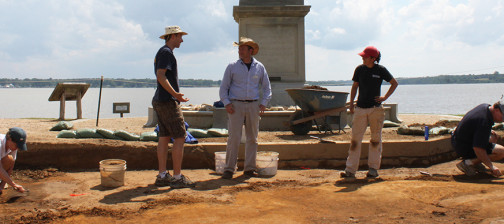 Three archaeologists stand in excavations in front of statue