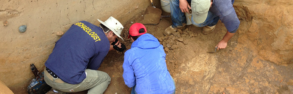 Archaeologist video recording excavations while others trowel nearby