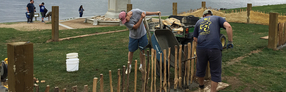 Men tip a wheelbarrow of dirt next to posts of a low wall