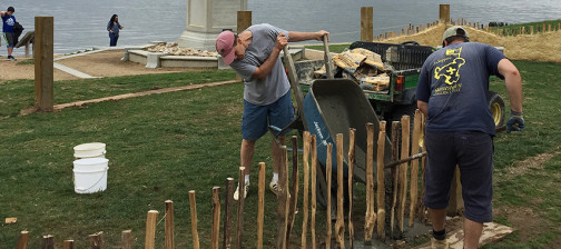 Men tip a wheelbarrow of dirt next to posts of a low wall