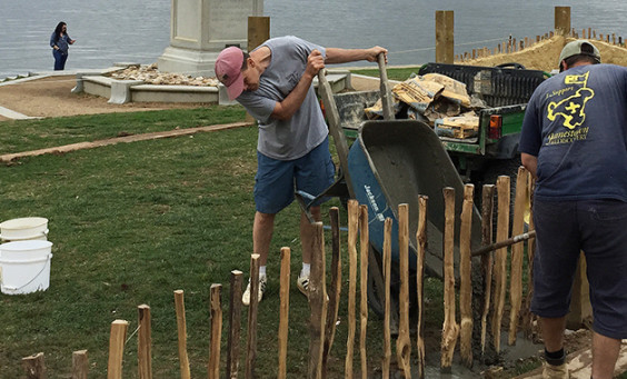 Men tip a wheelbarrow of dirt next to posts of a low wall