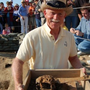 Archaeologist showing an excavated broadsword in a wooden tray to the camera while a group of visitors take photographs in the background