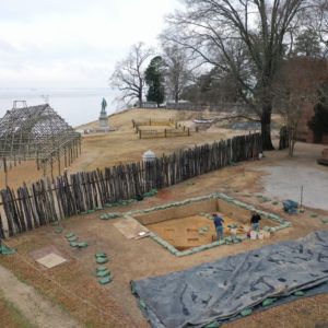 aerial view of two archaeologists shoveling in a large excavation unit next to a palisade and wooden building frame