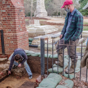 two men watch a woman pointing to a feature within an excavation unit