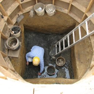 Archaeologist excavating a square well surrounded by a circular supporting wooden frame