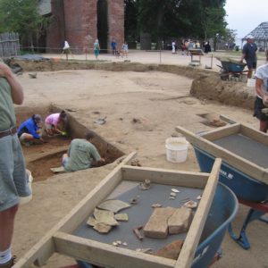 Screen of artifacts sitting on top of a wheelbarrow with archaeologists excavating and various groups walking in the background