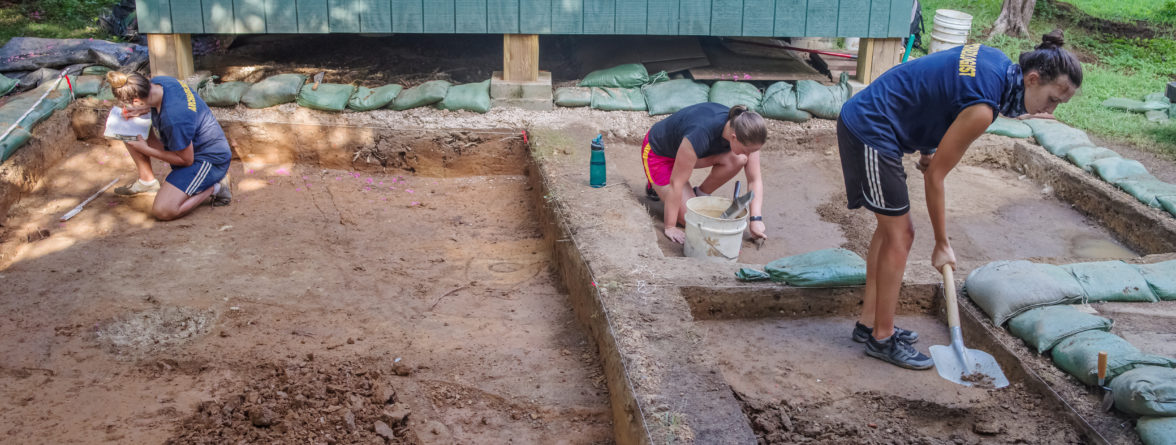 three archaeologists excavating in front of a wooden structure