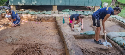 three archaeologists excavating in front of a wooden structure