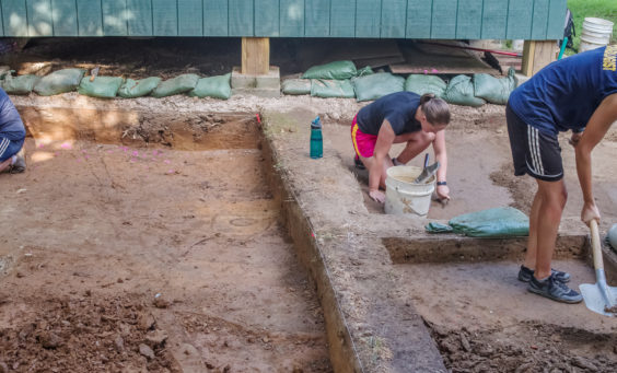 three archaeologists excavating in front of a wooden structure