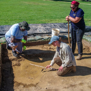 Aaron McKee of the grounds and maintenance team watches Staff Archaeologist Natalie Reid score a burial found south of the Archaearium on a staff dig day. Archaeological Field Technician Hannah Barch looks on.