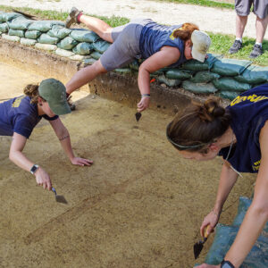 Archaeological Field Technicians Ren Willis, Hannah Barch, and Site Supervisor Anna Shackelford score the features in the burial area south of the Archaearium. They're preparing the area for record photography.