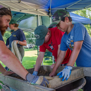 Archaeological Field Technician Josh Barber helps two campers look for artifacts while screening soil from the cellar site.