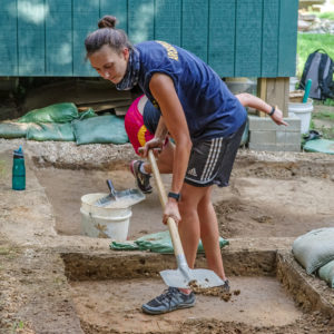 archaeologist shoveling dirt in an excavation unit in front of a wooden structure