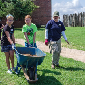 Two campers push a wheelbarrow of screened soil towards the discard pile with an assist from archaeological intern Faith.
