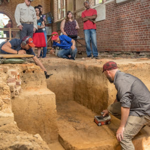 Man runs mini GPR unit across excavated feature while crowd watches