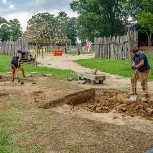 two archaeologists shoveling in excavation units in front of a reconstructed wooden structure and palisade walls