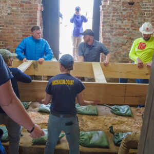 The archaeological team and contractors from Daniel and Company lower the base of the protective barrier that will shield archaeological resources during the concrete pour of the glass floor footings.