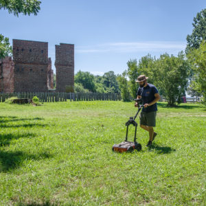 man pushing a ground-penetrating radar unit along a transect line in a field in front of brick ruins