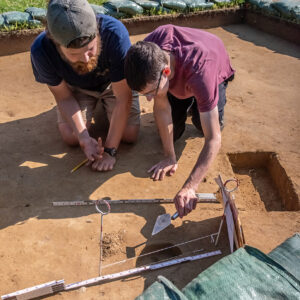 Archaeological Field Technician Josh Barber teaches Field School student Drew Parker how to bisect a posthole.