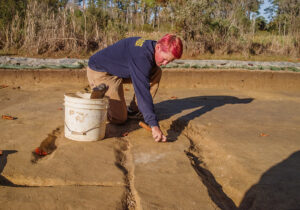 Staff Archaeologist Caitlin Delmas excavating at the borrow pit. Three plow scars are visible running vertically in the photo.