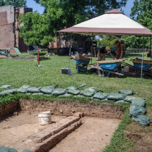 Open excavation unit and archaeologists screening artifacts under a tent