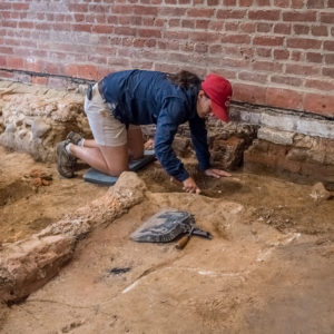 Kneeling archaeologist excavates dirt floor in front of brick wall